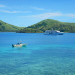 A small motorboat and a large cruise ship in blue water with green hills in the background