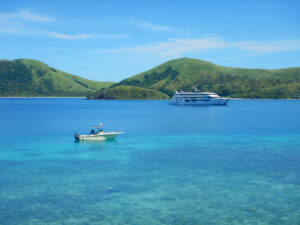 A small motorboat and a large cruise ship in blue water with green hills in the background