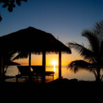 Silhouette of a cabana and palm tree by the water at sunset