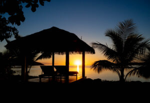 Silhouette of a cabana and palm tree by the water at sunset