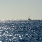 Distant sailboat crosses the horizon against a sparkling blue sea